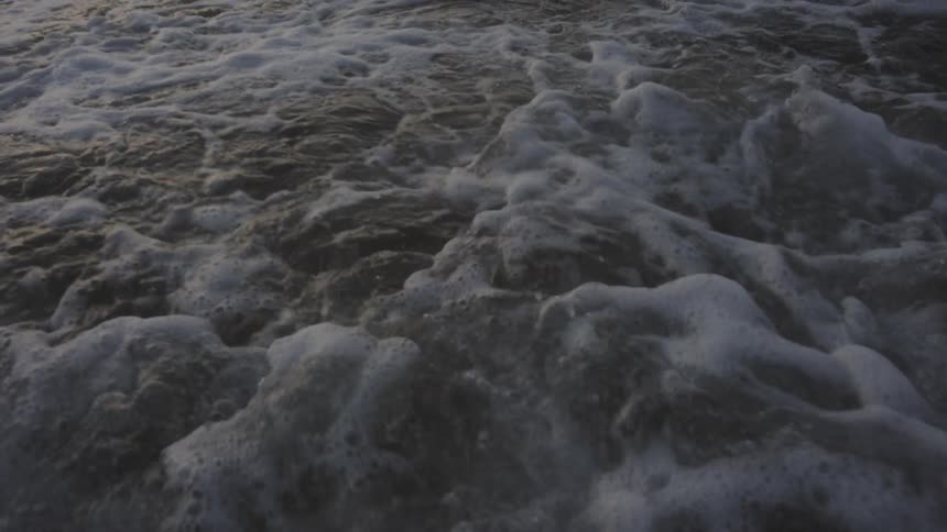The video provides a closeup shot of ocean waves rolling onto a sandy shore the foamy white crests of the waves crash and dissipate as they move toward the camera