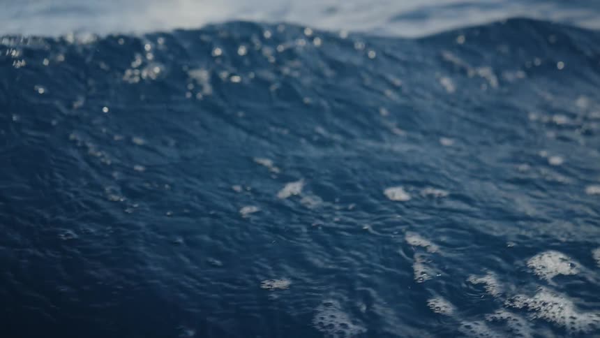A closeup view of the surface of the ocean shows small dark blue waves rippling and cresting the movement of the water is steady with patches of foam visible on the surface