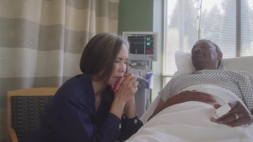 Woman sits beside a hospital bed patient holding her hands near her face