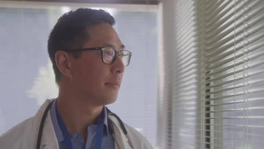 Doctor with a stethoscope around his neck looks out the window with blinds