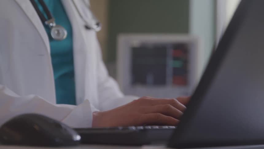 A shot of a medical professionals hands typing on a laptop computer the person is wearing a white coat and a stethoscope is visible around their neck
