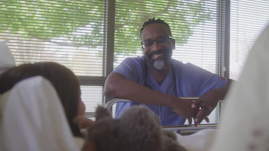 A male medical doctor sits beside a child in a clinic at midday wearing glasses a blue coat and a stethoscope around his neck smiling at the child