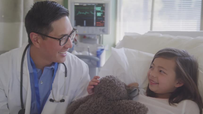 A male doctor smiles warmly at a young girl in a hospital bed holding a stuffed animal