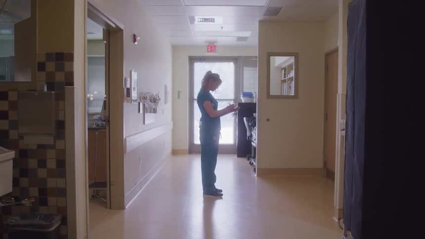 A female nurse stands in a hallway reviewing a clipboard in a softly lit hospital