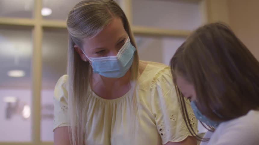 Woman smiles at a girl while they both wear face masks indoors