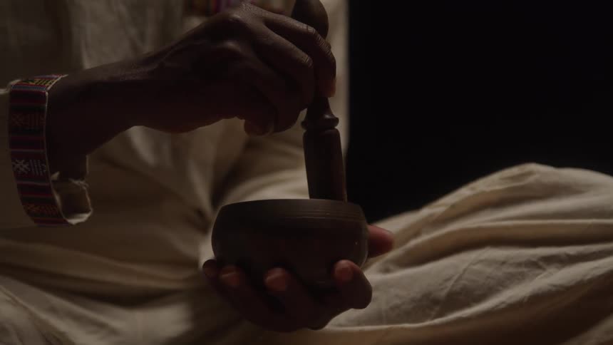 A person holds and plays a small wooden or metal singing bowl the person is wearing lightcolored clothes and their hands and the bowl are silhouetted against a dark background
