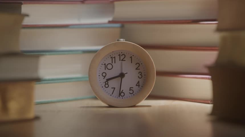 Clock hands move around the face of an analog clock standing between stacks of books