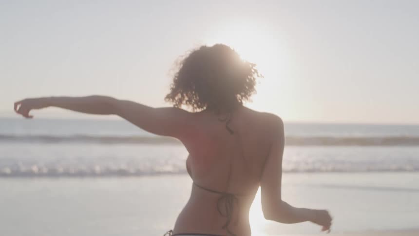 Woman in a gray bikini stretching on the beach with the sun in the background