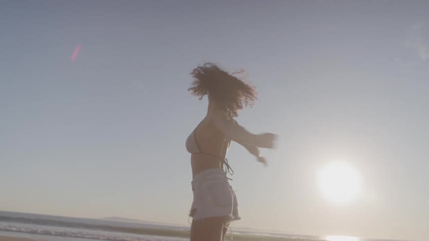 A woman with curly dark hair wearing a dark bikini top and a pair of light colored shorts is standing on a sandy beach at sunset as she smiles spins around with her arms in the air and looks up at the sky