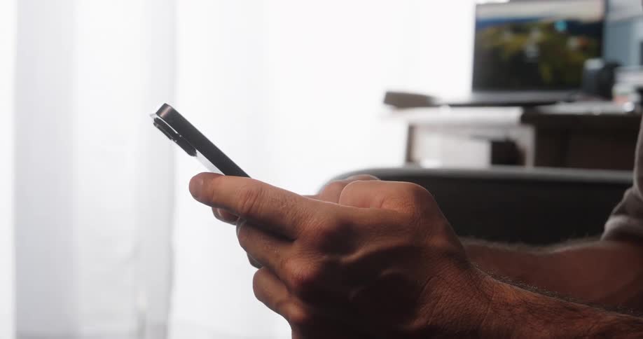 A persons hands are shown holding and typing on a smartphone with a laptop and bright background out of focus behind them