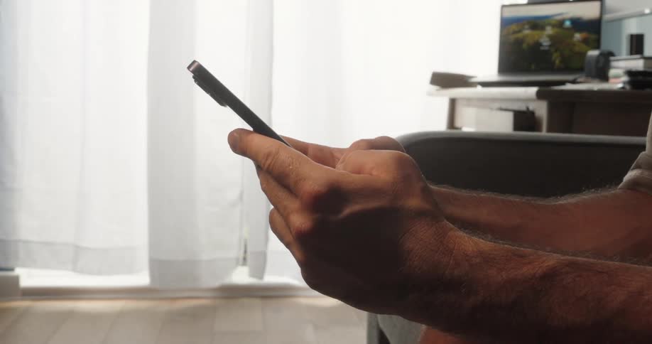 A mans hands are holding a smartphone with a blurred background of a room that includes a window with white curtains and a desk with a laptop