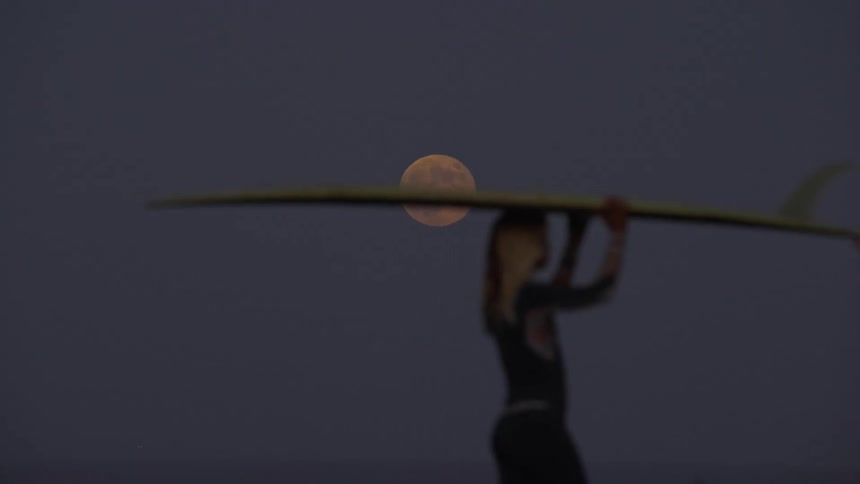 Surfer woman walking and carrying her surfboard with a big fin under the full moon light at night