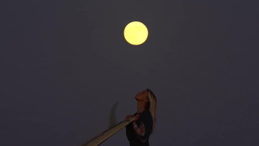 Surfer woman walking and carrying her surfboard with a big fin under the full moon light at night