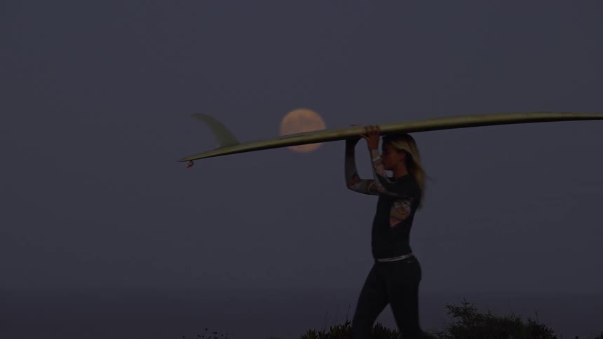 Surfer woman walking and carrying her surfboard with a big fin under the full moon light at night