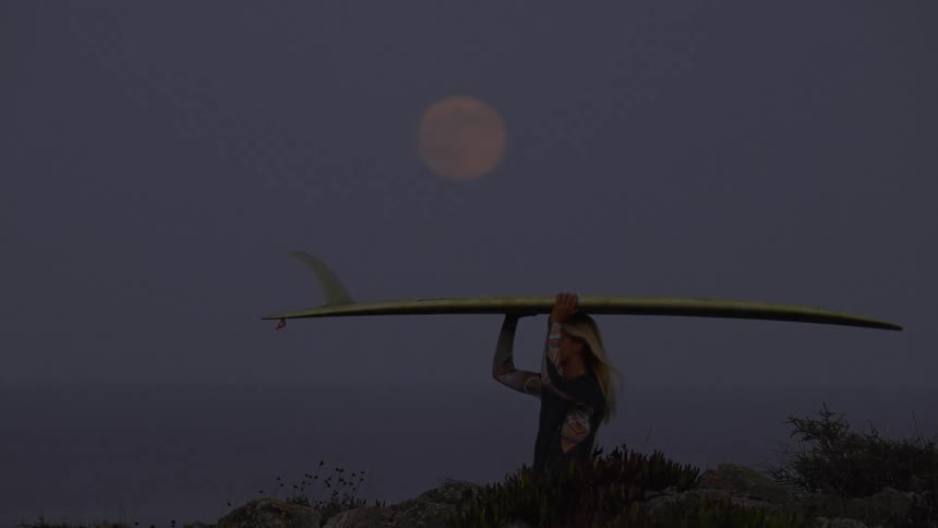 Surfer woman walking and carrying her surfboard with a big fin under the full moon light at night
