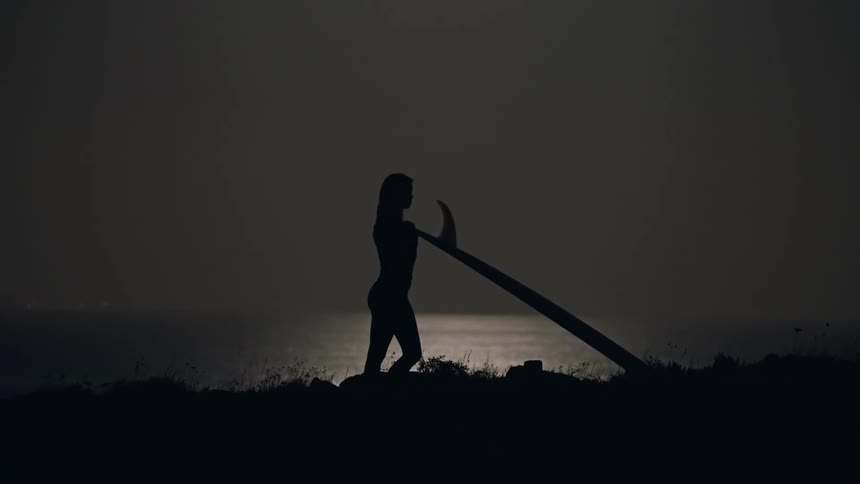 Silhouette of a woman standing on a cliff next to the sea holding a surfboard at night under full moon light