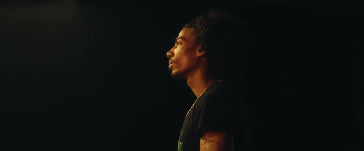 A young man stands in a dark room wearing a black tshirt a necklace and a tattoo on his arm