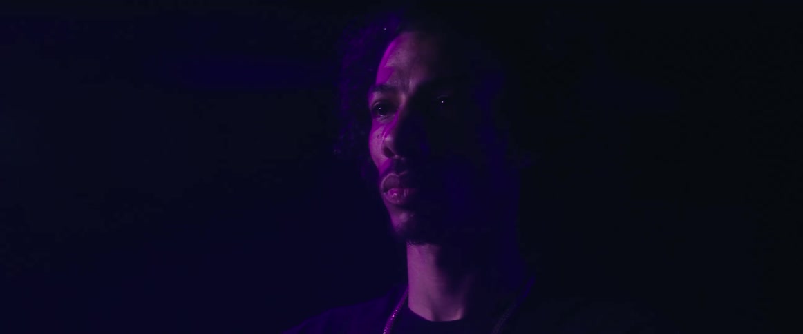 A man with curly hair is illuminated from the side by a shifting purple light in a dark setting