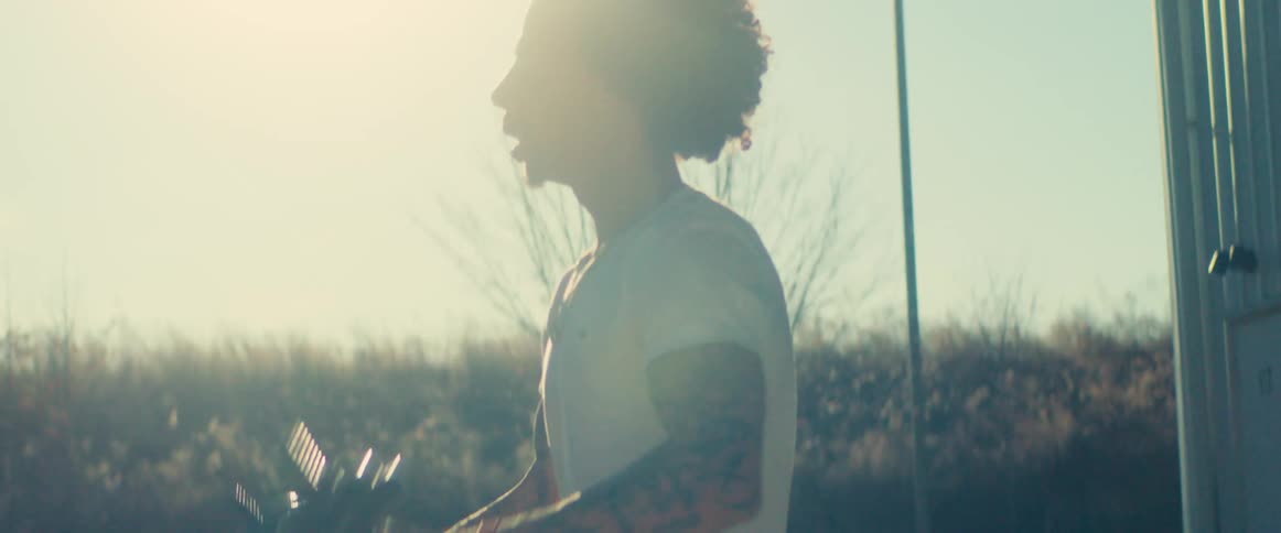A man with dark curly hair and tattoos on his arms wearing a white shirt is standing outside and doing an exercise lifting dumbbells