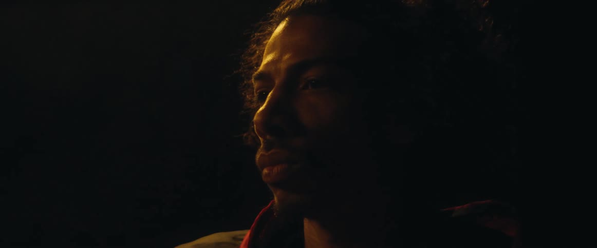 A man with dark curly hair is looking off to the left with a serious expression as he is lit from the side at night