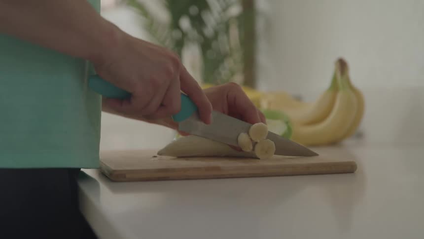 A persons hands using a knife to slice a banana on a wooden cutting board with more bananas and a plant in the blurry background