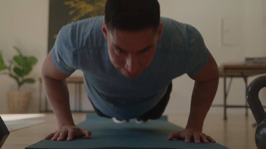 A man wearing a blue shirt and black shorts is on a blue yoga mat on a wooden floor doing pushups while a small kettlebell and a plant are visible in the room