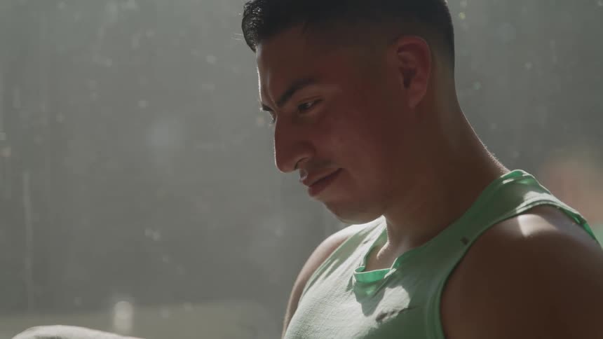 A man with dark hair wearing a green tank top is looking down with a focused and relaxed expression on his face near a sunlit window