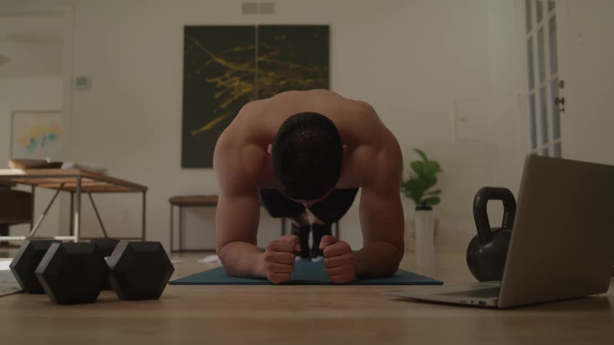 A young man barechested and wearing only trunks does a plank workout on a yoga mat in his living room at midday