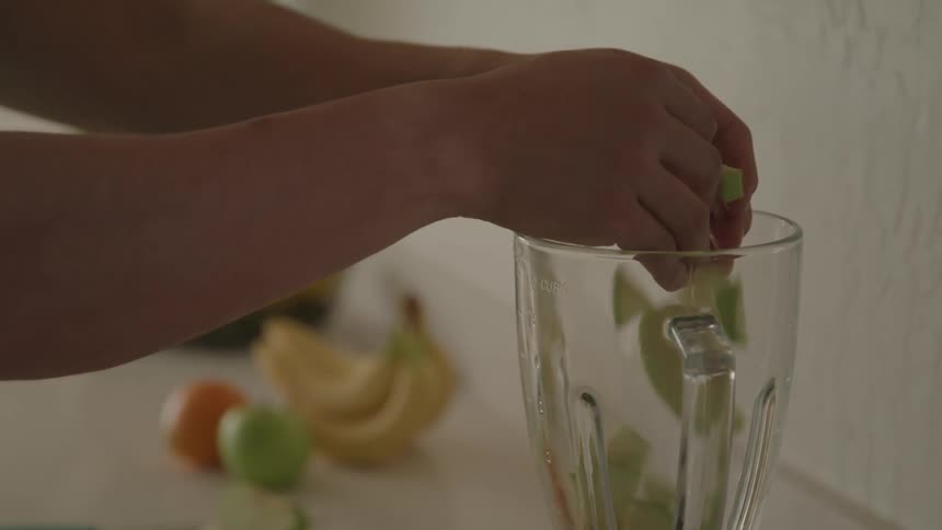 A persons hand places slices of apple into a blender on the countertop with other fruits lying beside it at midday