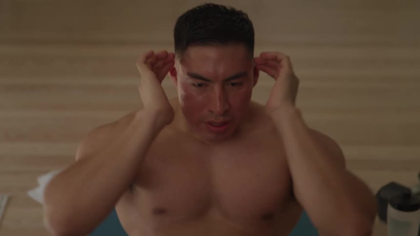 A shirtless young man does situps on a mat on the floor at midday