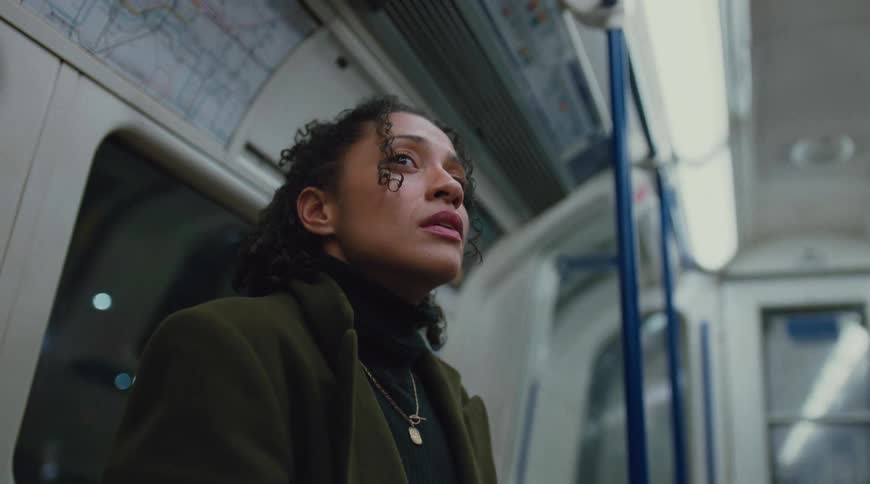 A young woman with curly hair wearing a green overcoat and a necklace sits in a train during a ride staring sadly at something beside her