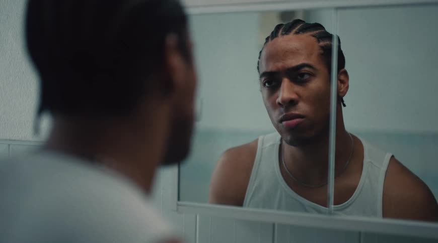 A young black man with braids washes his face with water in a bathroom sink in front of a mirror angrily staring at himself while wearing a singlet