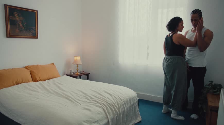 A woman sits on a bed and then stands up to embrace a man standing by the window