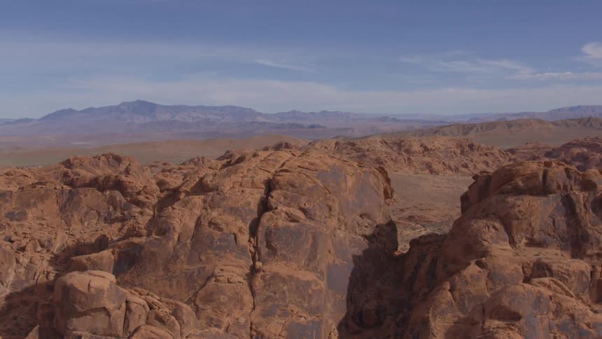 Sunlit reddishbrown rock formations stretch across the foreground with distant desert plains and hazy mountains under a clear blue sky