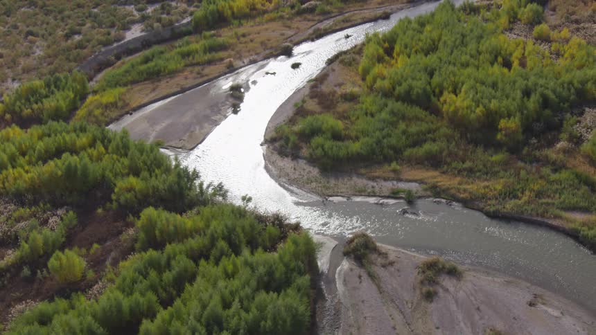 An aerial view shows a meandering river winding through a lush verdant landscape the sunlight glints off the waters surface as the camera pans along the river revealing dense groves of trees with patches of vibrant yellow and deep green foliage