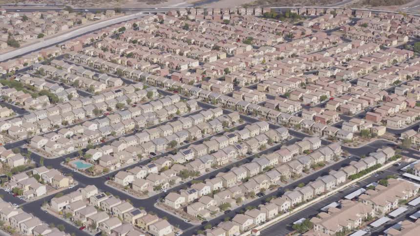 An aerial shot shows a vast uniform suburban landscape under a bright sunny sky rows upon rows of identical beige houses stretch out in every direction interrupted only by the gridlike pattern of streets