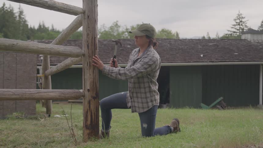 A woman dressed in a plaid shirt and a baseball cap kneels on a grassy lawn next to a wooden fence she is focused on hammering a nail into the fence post with a rustic barn visible in the background