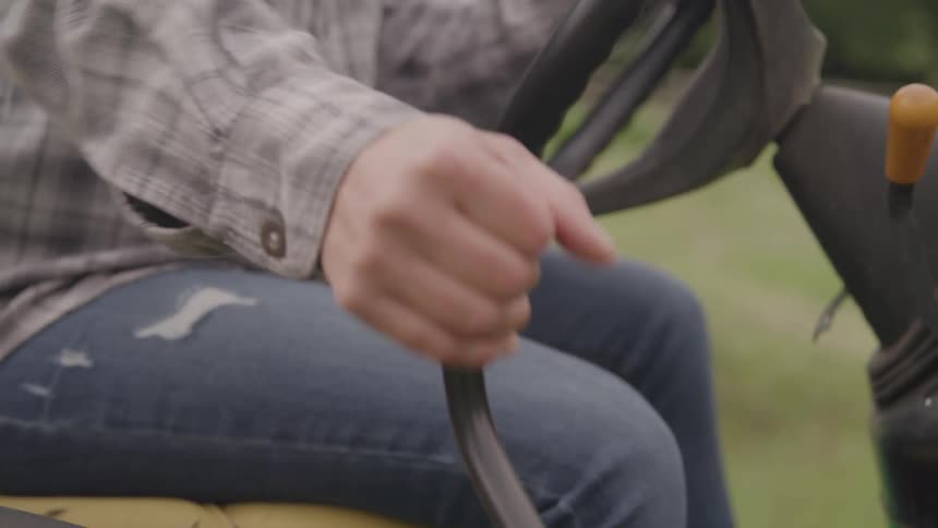 A closeup shot focuses on a persons hands gripping the steering wheel of a tractor the person is wearing a gray plaid shirt and jeans and the camera highlights the control levers and yellow seat of the vehicle as they prepare to drive