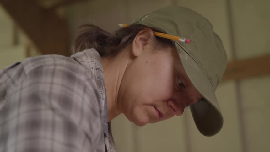 A closeup shot focuses on a persons face as they work indoors a pencil tucked behind their ear they are wearing a plaid shirt and a baseball cap looking down intently as they concentrate on a task out of frame