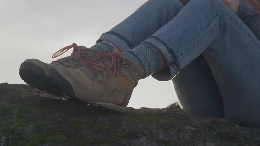 A closeup shot focuses on a persons legs and feet showing them wearing hiking boots with red laces and wool socks and blue jeans with rolledup cuffs the person is sitting on a rocky surface with the sun low in the sky behind them