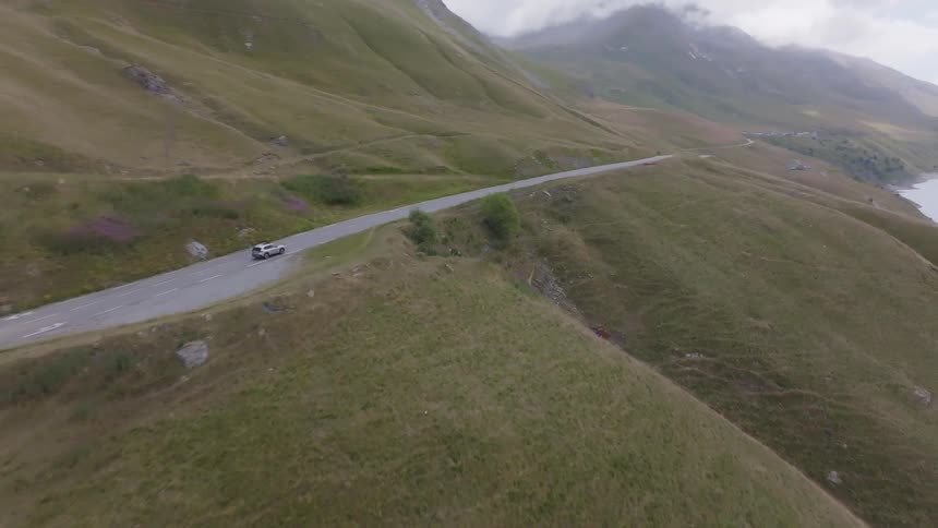 A highangle drone shot shows a white car driving along a winding mountain road the road is surrounded by rolling hills covered in brown and green grass with clouds looming in the distance
