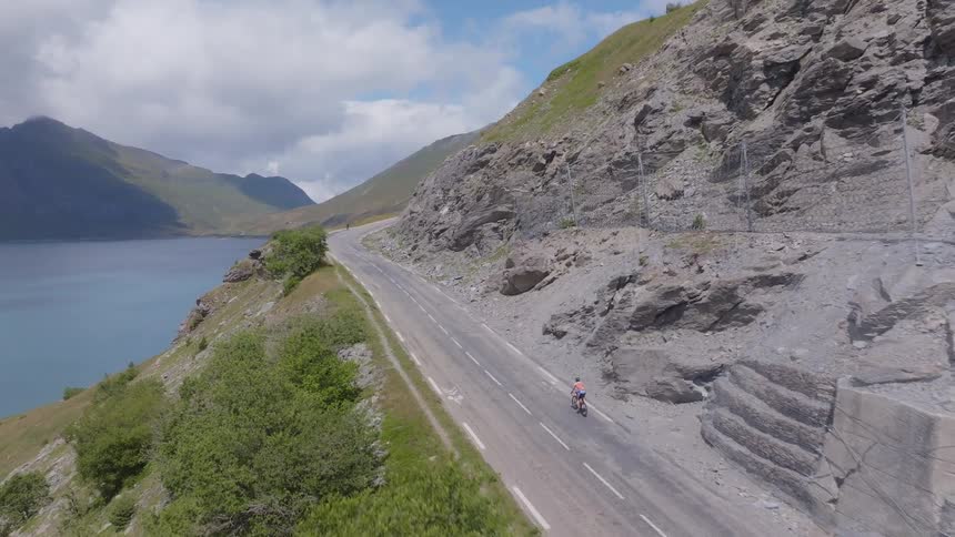 Cyclist rides along a coastal road beside a blue lake and rocky mountain