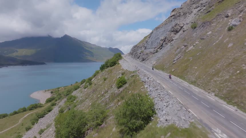 An aerial view shows two motorcycles driving along a winding mountain road that overlooks a large blue lake with mountains and a cloudy sky in the background