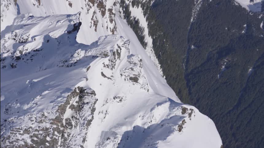 A jagged snowcovered mountain ridge rises sharply above dense green forest in the valley below