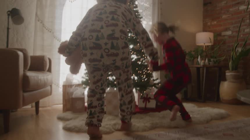 Two siblings run excitedly to the christmas tree in the living room hugging each other and wearing pajamas at midday