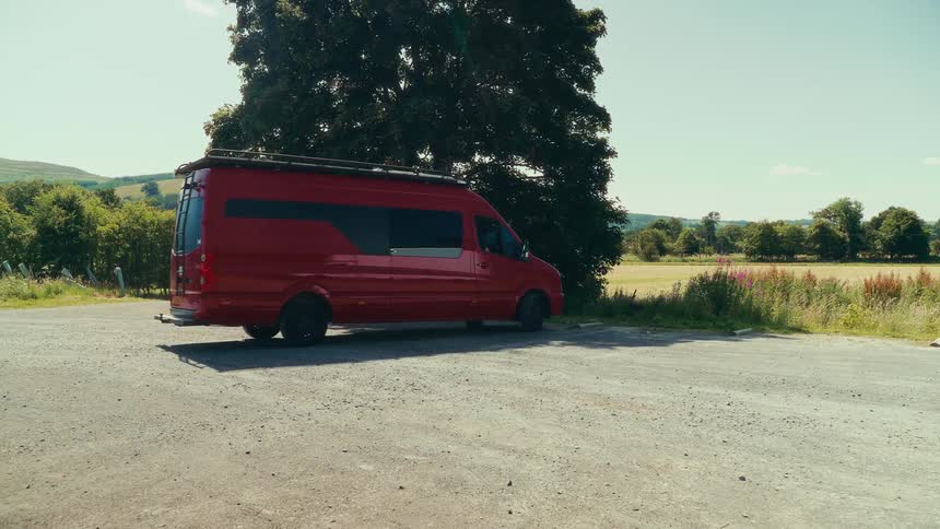 The video shows a wide shot of a red camper van that drives into a parking spot under a large tree the van stops and parks with its rear facing the camera the video has a watermark that reads filmsupply