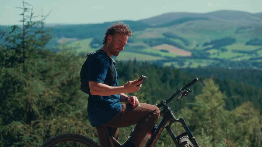 A man sitting on a mountain bike takes a selfie while flashing a peace sign against a backdrop of rolling green hills