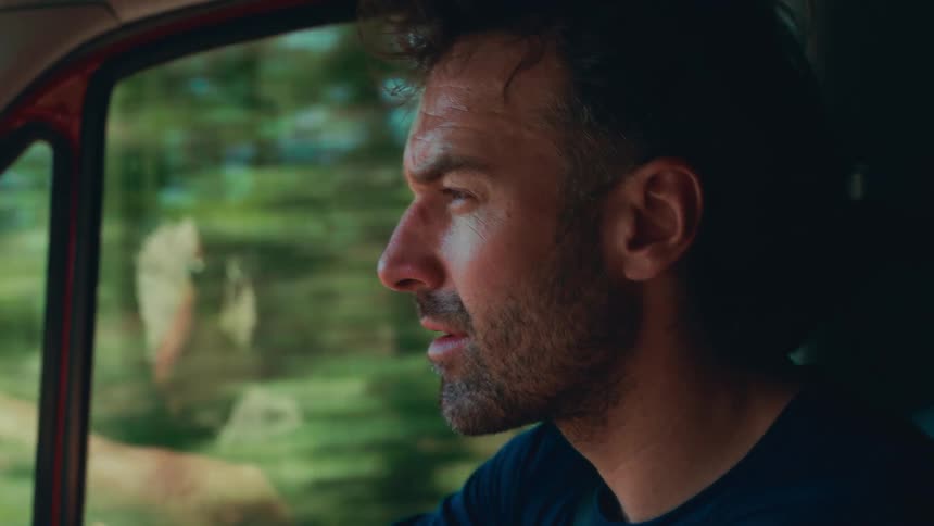 A man with short hair and a beard drives a vehicle looking intently at the road ahead through the open window