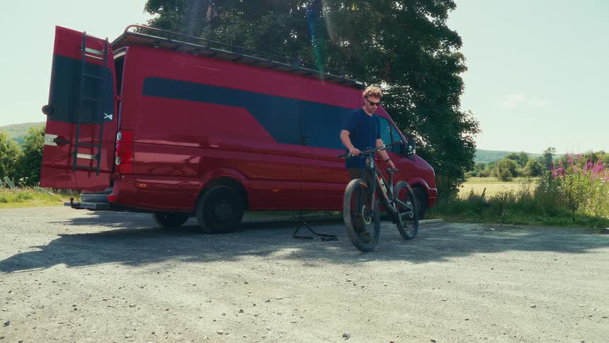 The video shows a man standing next to a mountain bike and a red camper van he picks up the bike and walks it past the van away from the camera the video has a watermark that reads filmsupply