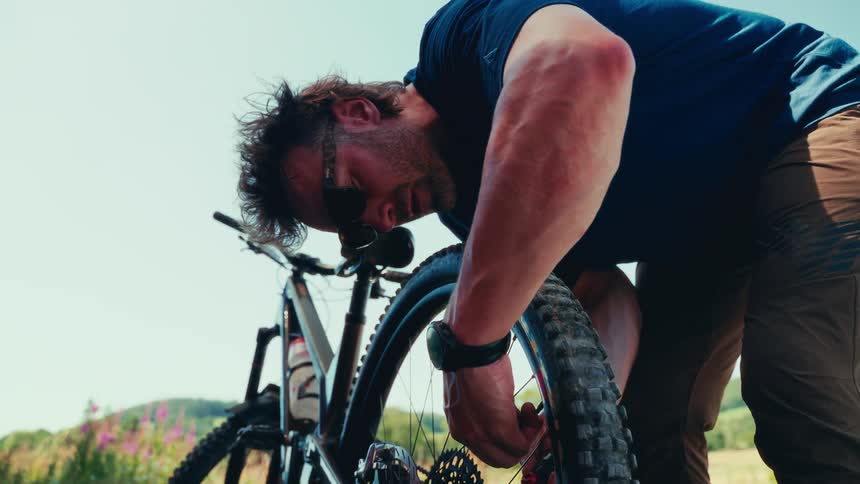A man in sunglasses closely inspects the tire of his mountain bike on a sunny day